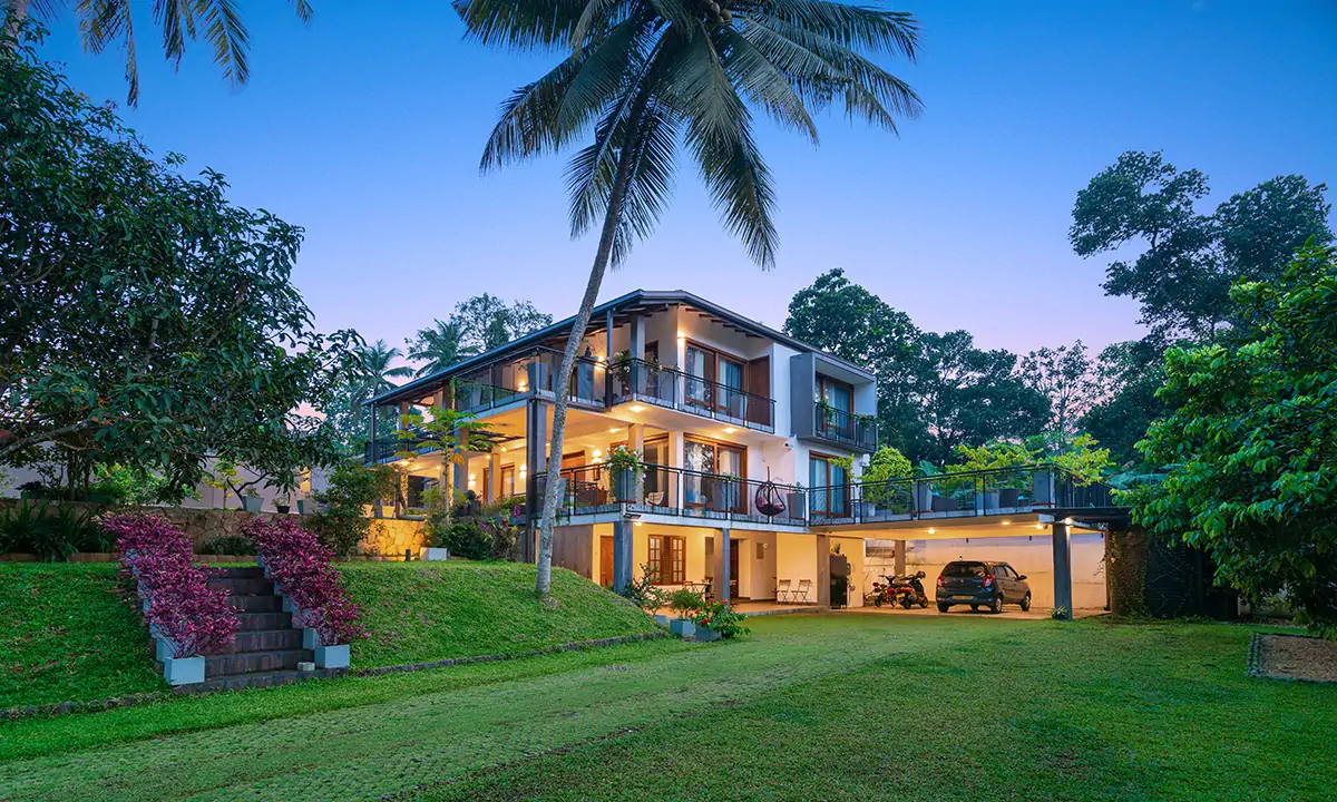 The house on its sloping garden at dusk, with steps up the lawn and coconut palms