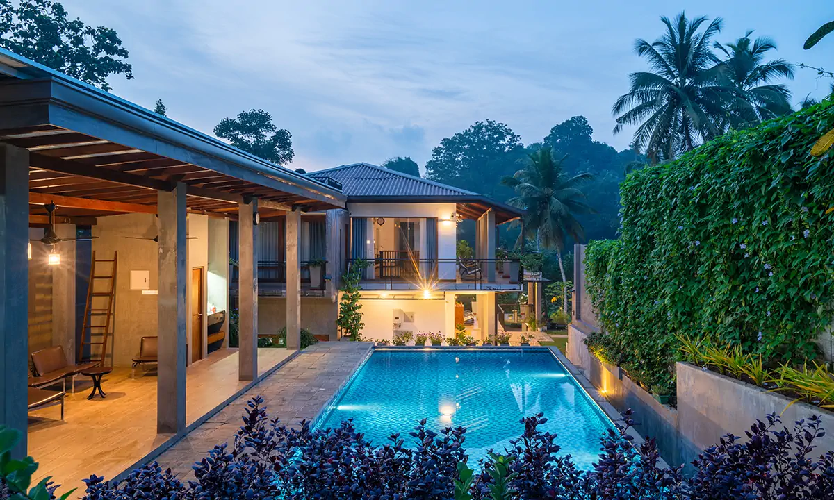 Swimming pool between timber-roofed pavilions and a planted wall, at dusk