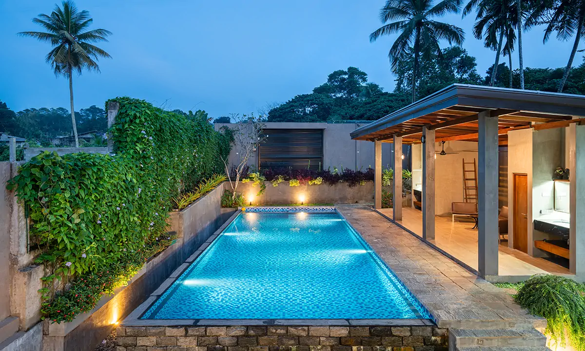 Pool lit at dusk between a green wall and a timber-roofed pavilion