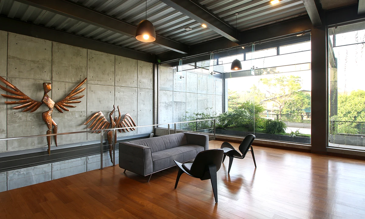 Lounge with a timber floor, a fair-faced concrete wall and a winged sculpture, glazed to the trees