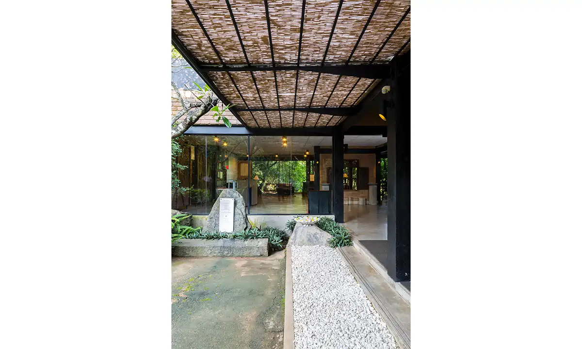 Covered entrance path beside eco-brick walls, under a shade screen
